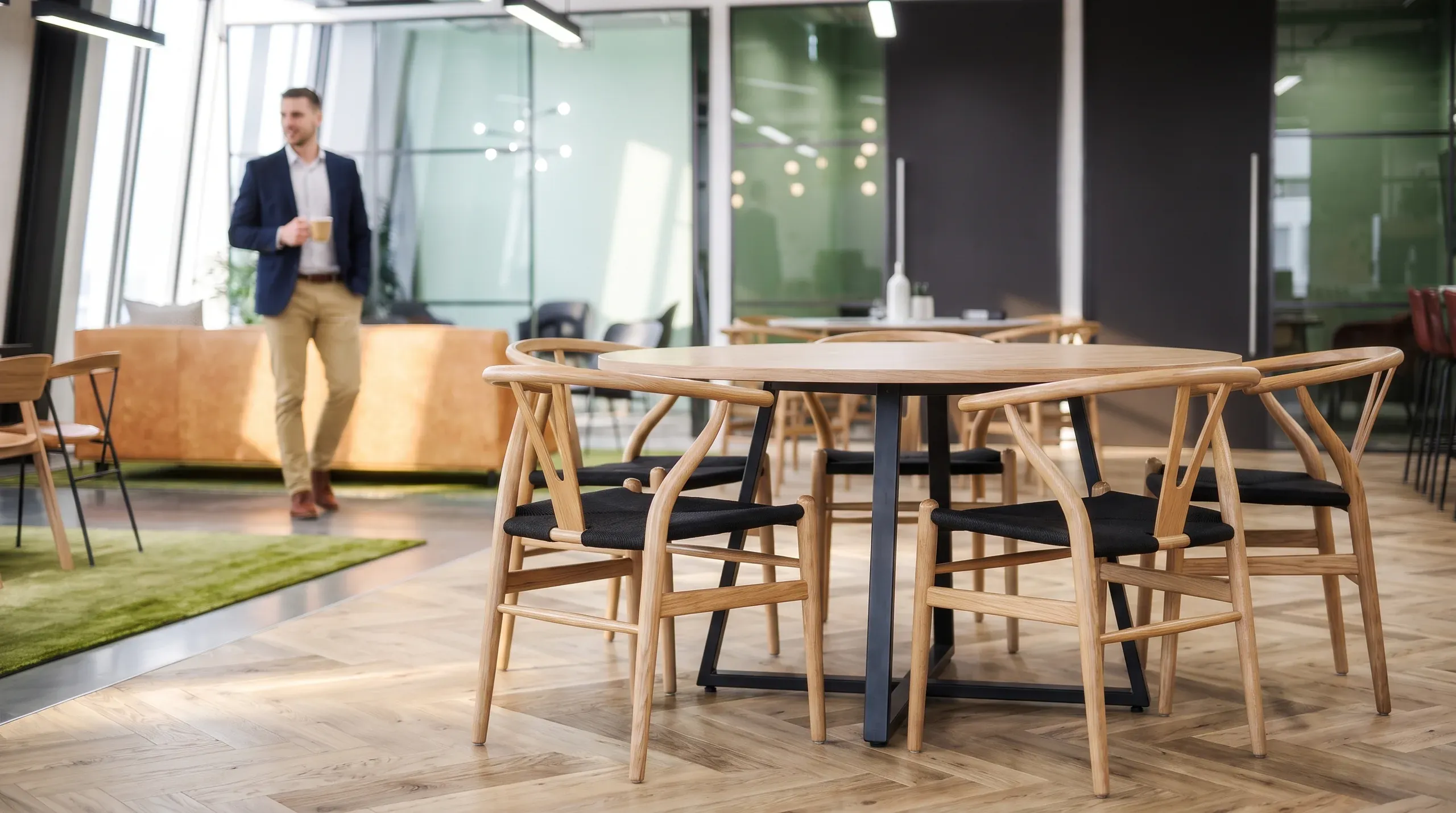 Modern office break room with wooden chairs around a table, man in business casual attire in background, glass partitions, green moss wall accent.