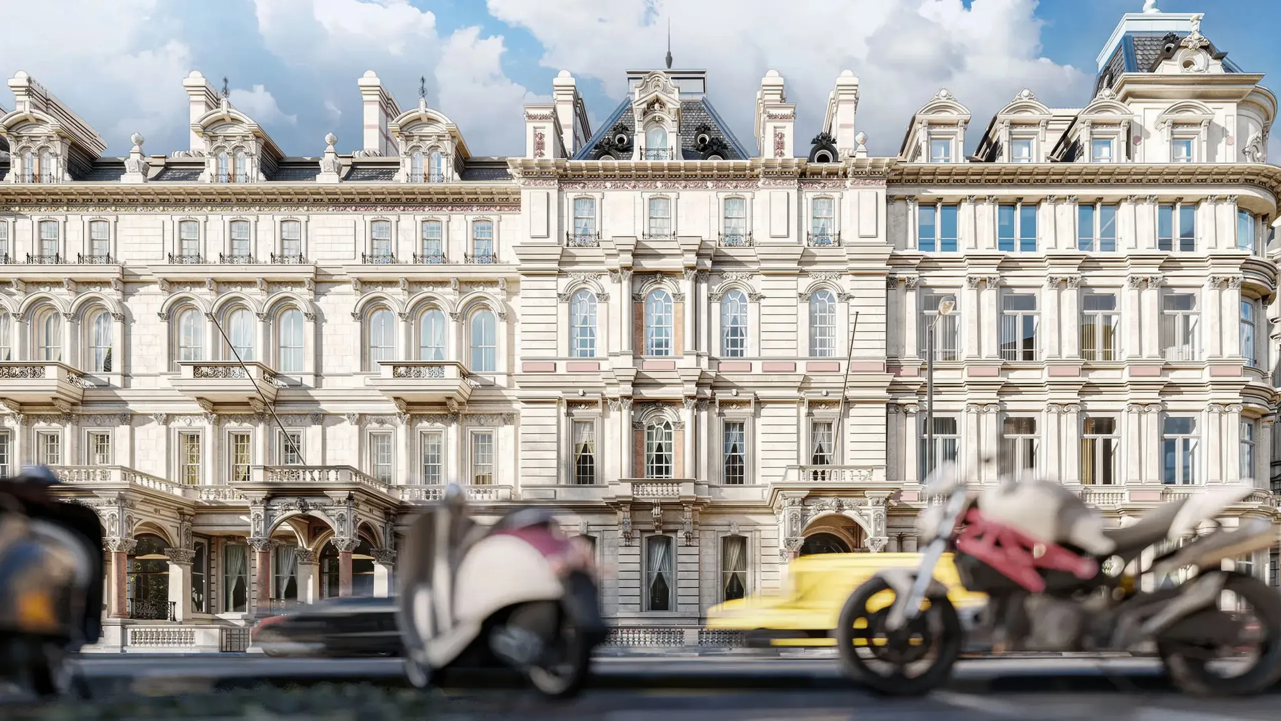 Luxury Parisian building facade with classical architecture, ornate mansard roof, and blurred vehicles passing in foreground under blue sky.
