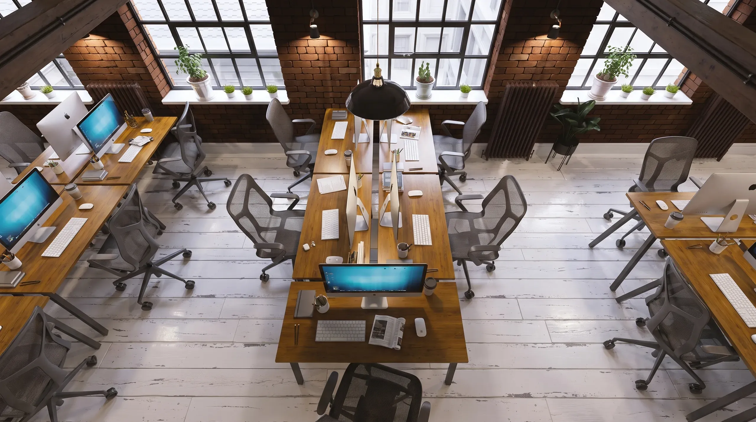 Modern loft office with wooden desks, gray mesh chairs, skylights, exposed brick, and potted plants along windows.