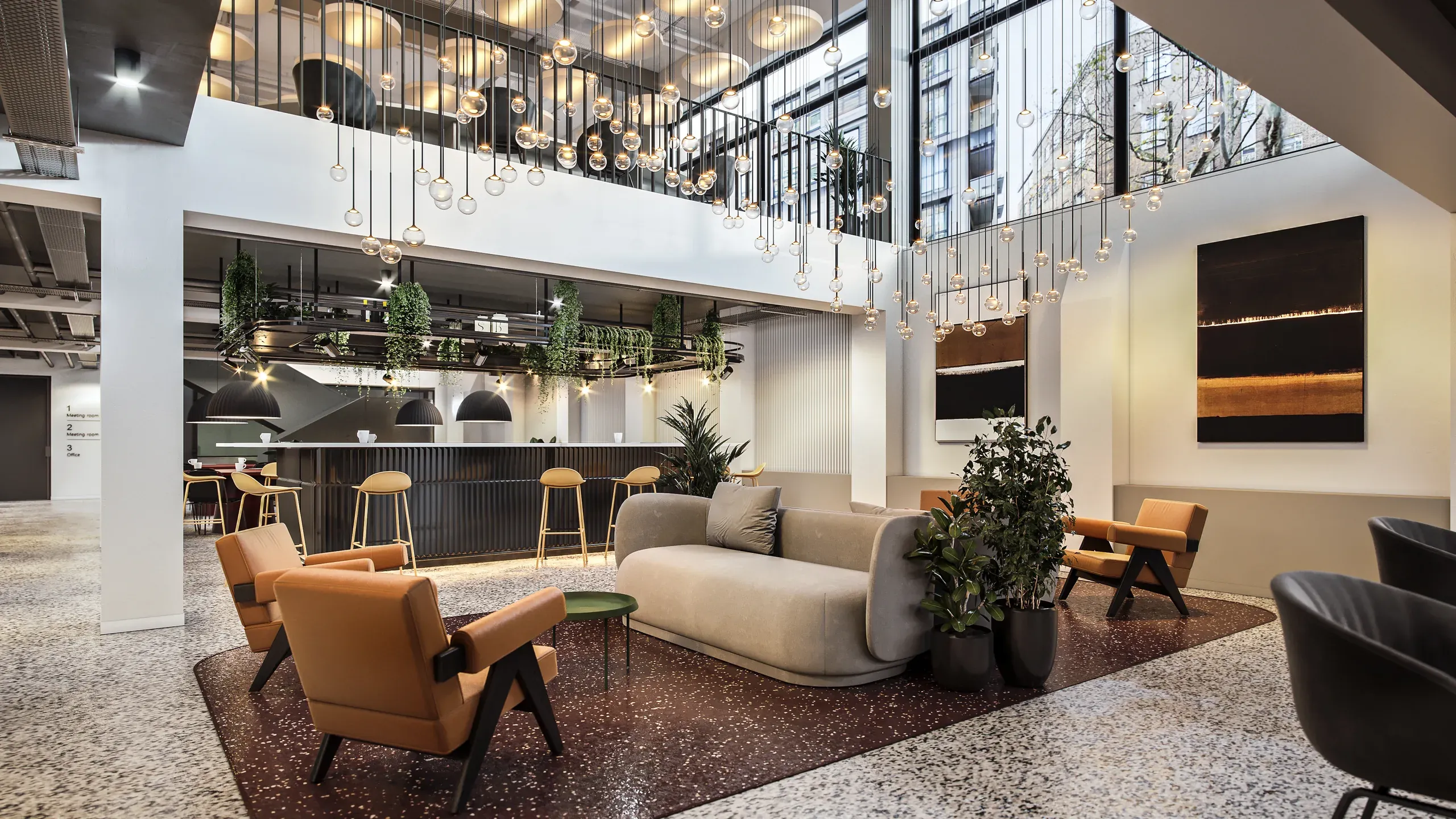 Modern hotel lobby with high ceilings, pendant lighting, orange accent chairs, cream sofa, bar seating, and floor-to-ceiling windows overlooking city buildings.