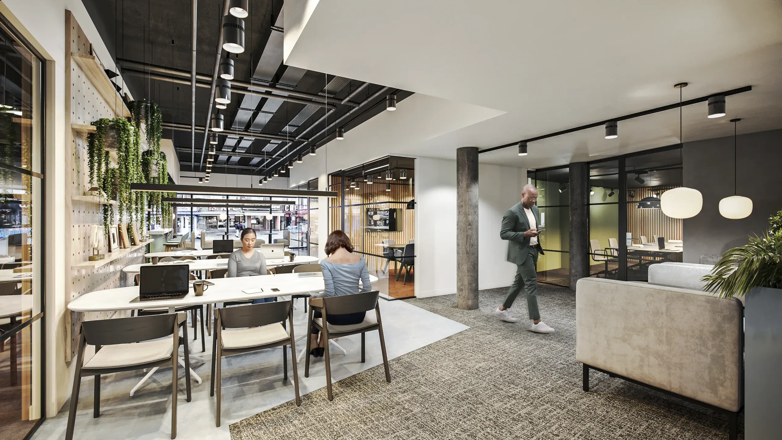 Modern open-plan office space with workers at collaborative desks, exposed ceiling with track lighting, concrete columns, and green plant accents.