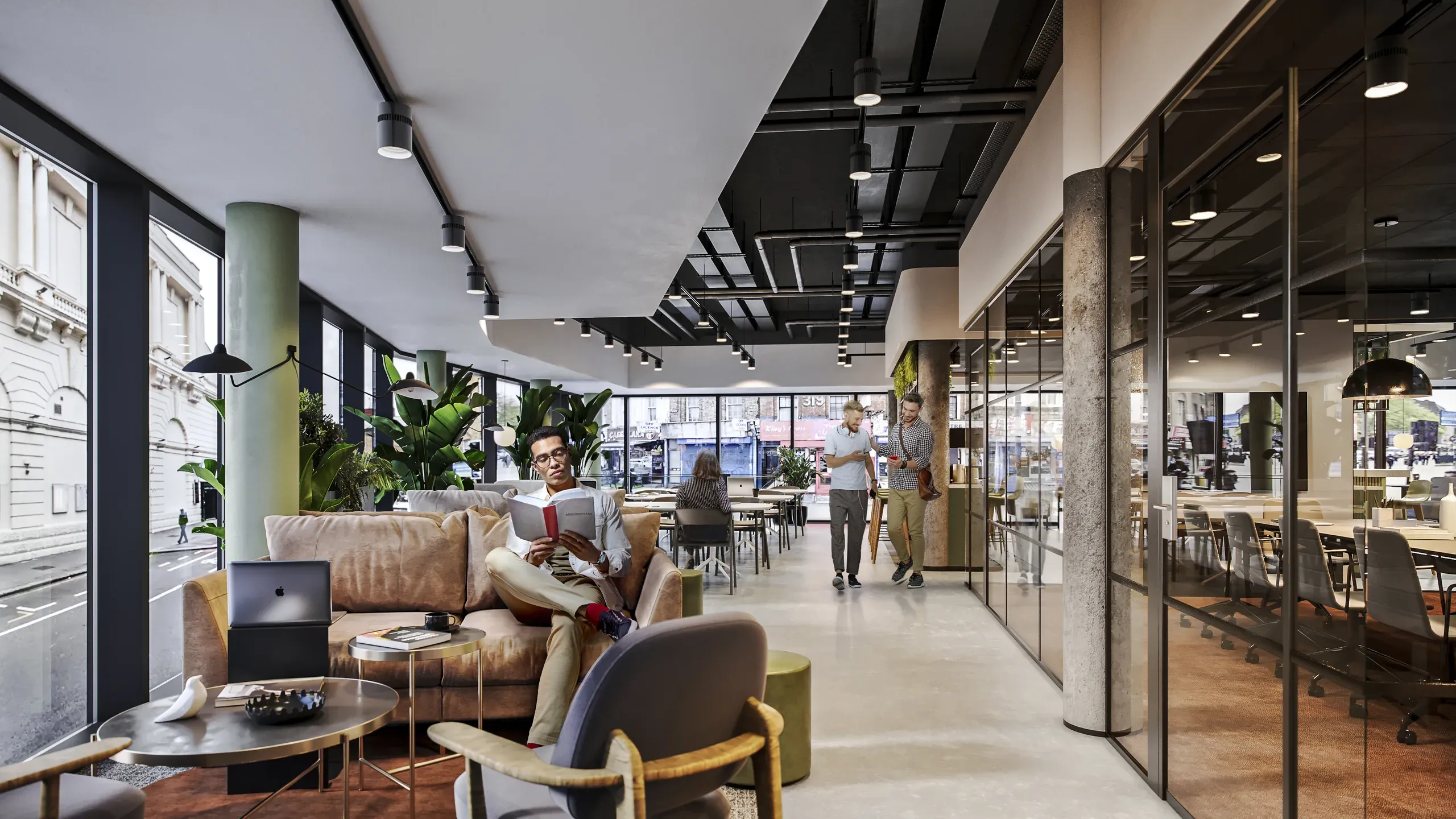 Modern open-plan office space with lounge seating, glass-walled meeting rooms, exposed columns, and large windows overlooking an urban street.