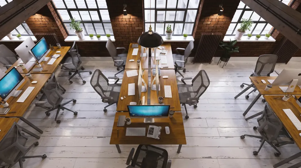 Modern loft office with wooden desks, gray mesh chairs, skylights, exposed brick, and potted plants along windows.