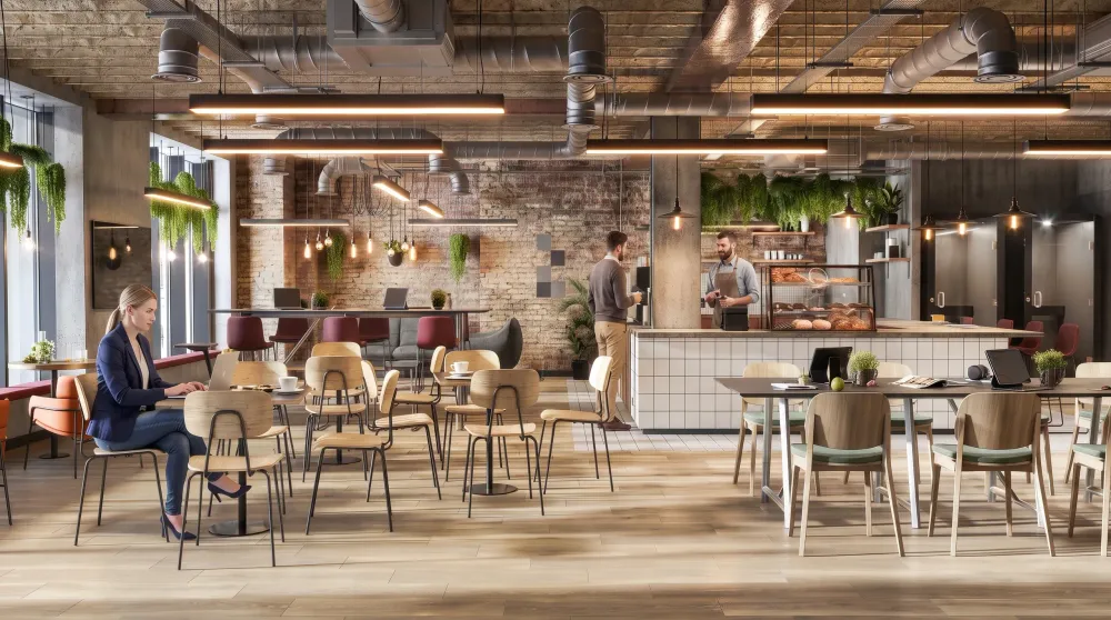 Modern office cafeteria with exposed brick walls, industrial ceiling, natural wood flooring, pendant lighting, green plants, and employees working at tables near a counter service area.