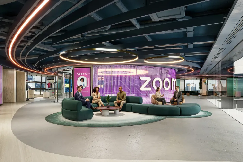 Modern Zoom office lobby with curved green seating, purple welcome signage, circular ceiling lights, and five people in business casual attire conversing.