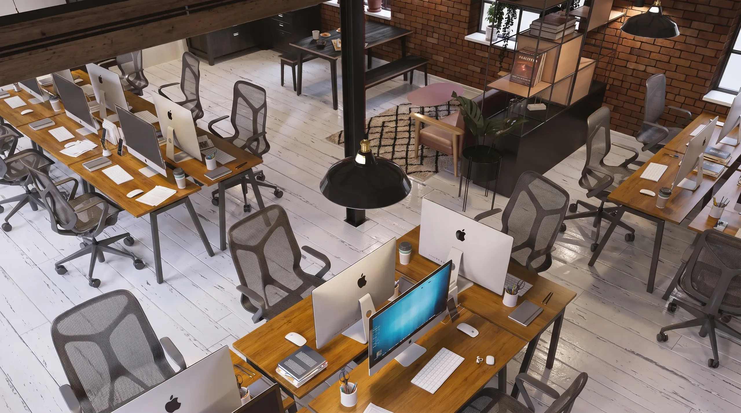 Modern open-plan office space with wooden desks, mesh chairs, Apple computers, brick walls, and a central pendant light fixture.