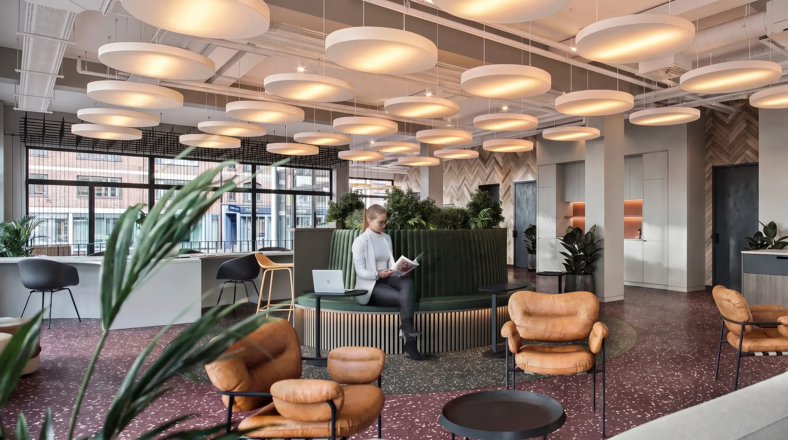Modern office lounge with pendant lighting, leather seating, green plants, and a woman working at a laptop by a green partition wall.