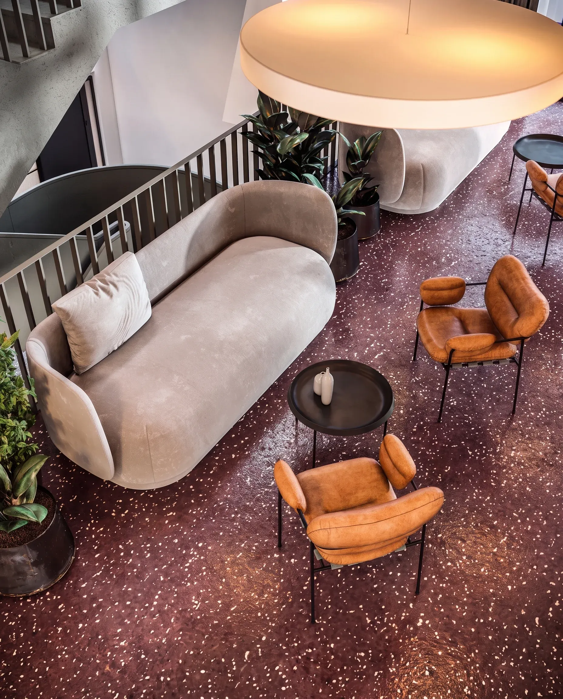 Modern lounge area with curved beige sofa, cognac leather chairs, black side table, potted plants, and pendant lighting overhead, shot from above.