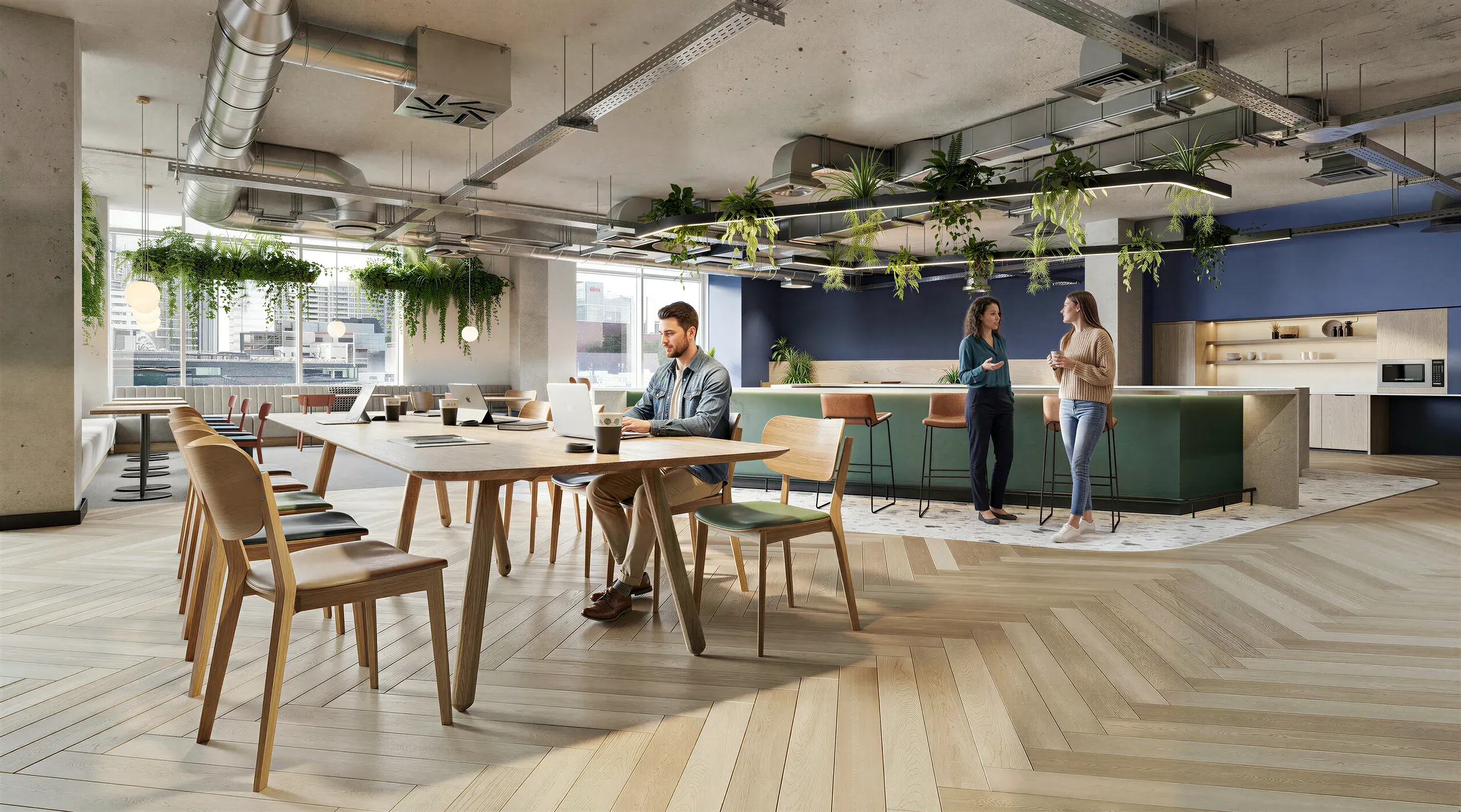 Modern office break room with wooden tables, tan chairs, hanging plants, concrete ceiling, blue accent wall, and employees socializing by a green counter kitchen area.