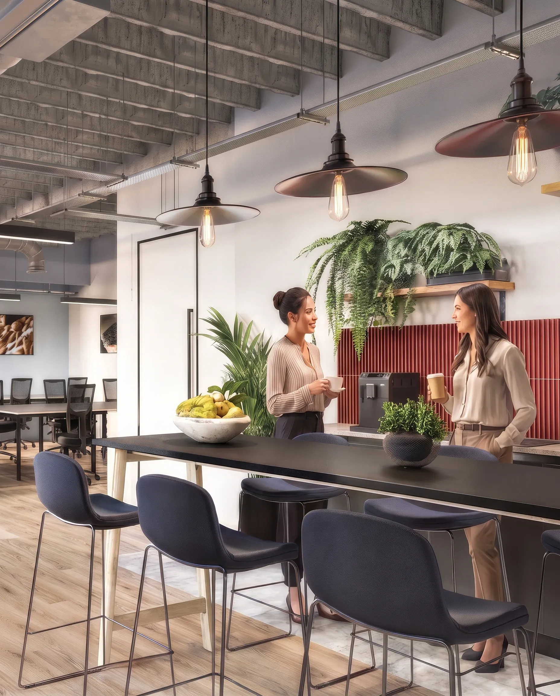 Modern office break room with two women chatting at a black counter, pendant lights overhead, potted plants, and open workspace visible in background.