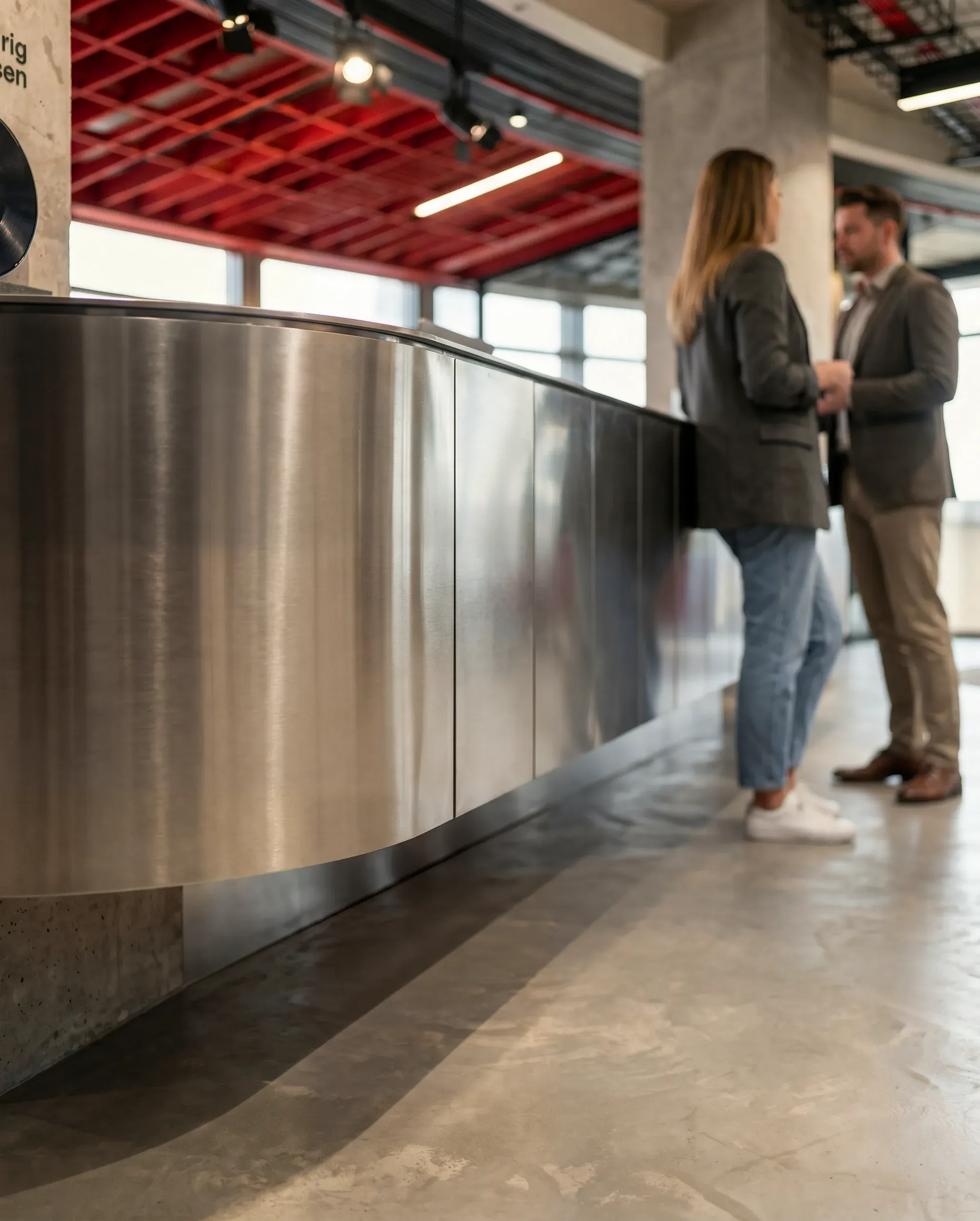 Two people in business attire stand in a modern interior with curved stainless steel counter and red ceiling panels with ambient lighting.
