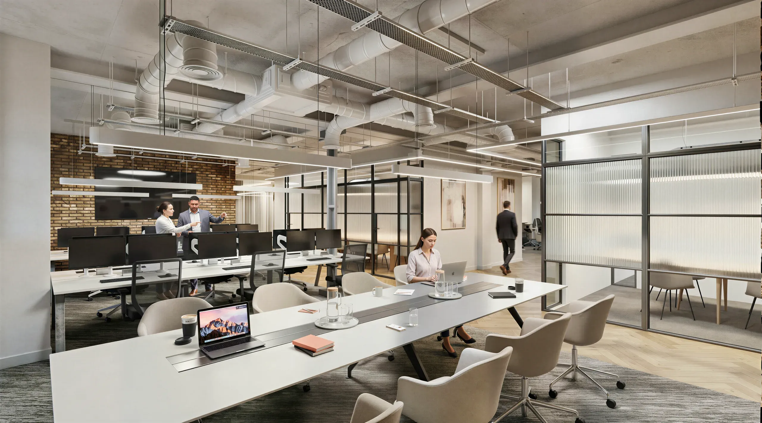 Modern open-plan office space with glass partitions, collaborative work areas, exposed ceiling ducts, brick accent wall, and multiple employees working at desks and meeting tables.