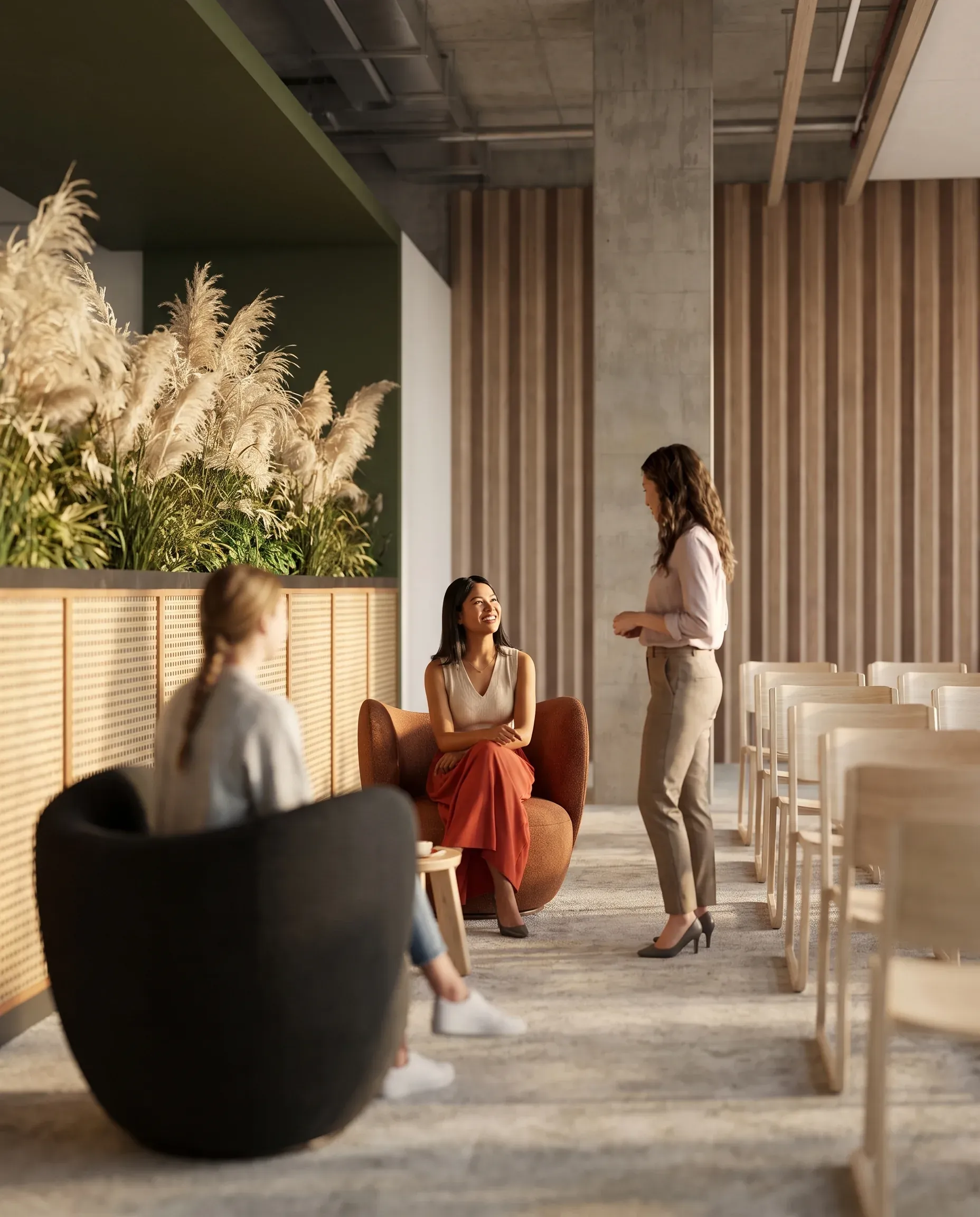 Modern office lounge with three women conversing, featuring contemporary furnishings, pampas grass décor, and minimalist architectural design with concrete columns.