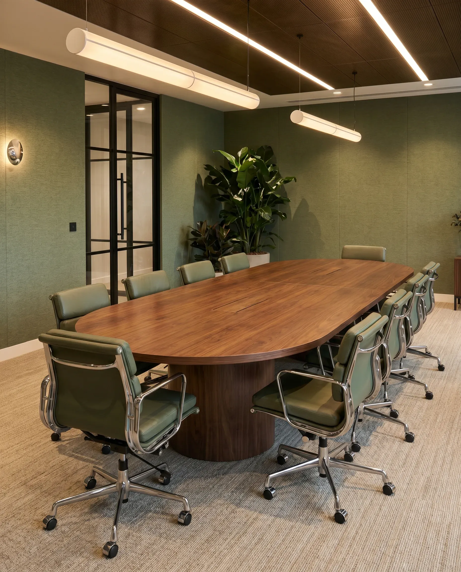 Modern conference room with oval wooden table, green leather chairs, sage green walls, pendant lighting, and potted plant by glass door.