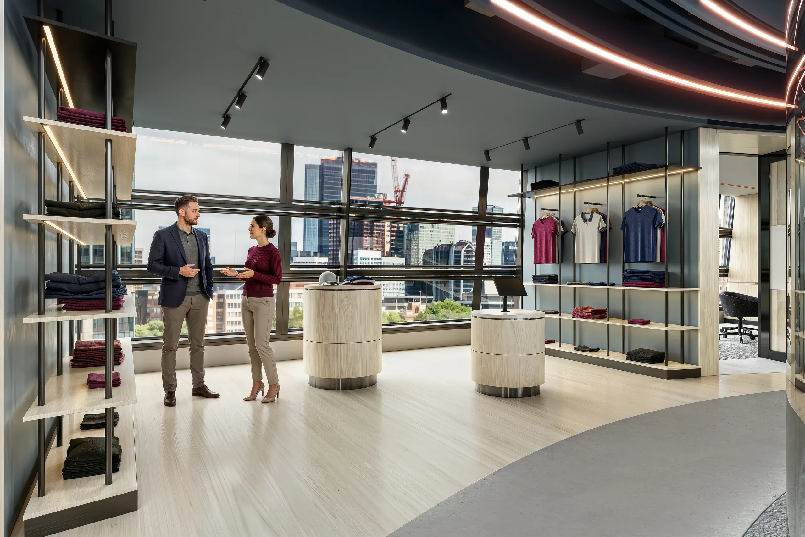 Modern office showroom with two professionals conversing near cylindrical display units, clothing racks, shelving, and floor-to-ceiling windows overlooking city skyline.